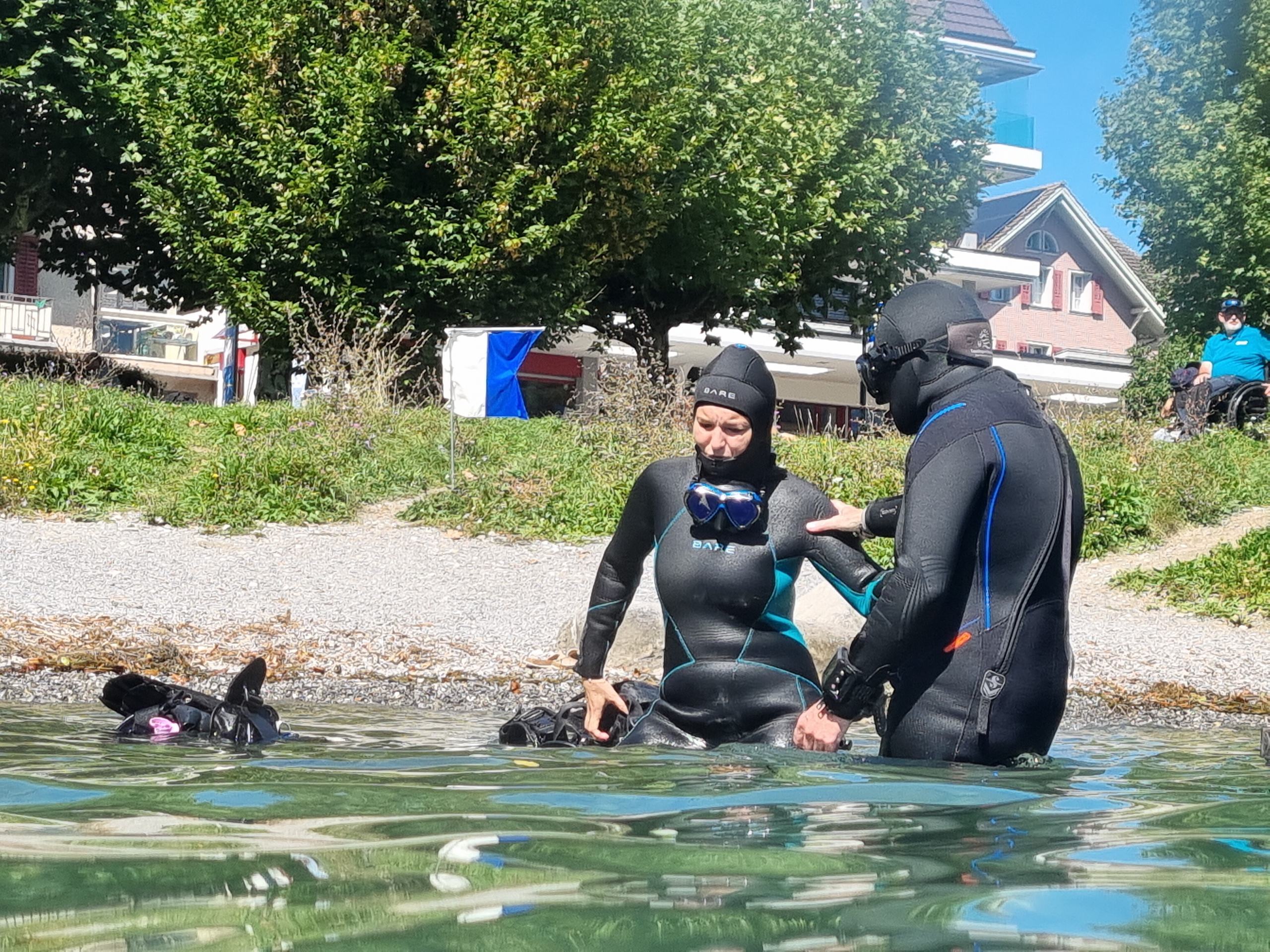 Zwei Menschen im Neoprenanzug im flachen Wasser des Vierwaldstättersees am Hausriff Hergiswil, die Instruktorin erklärt.