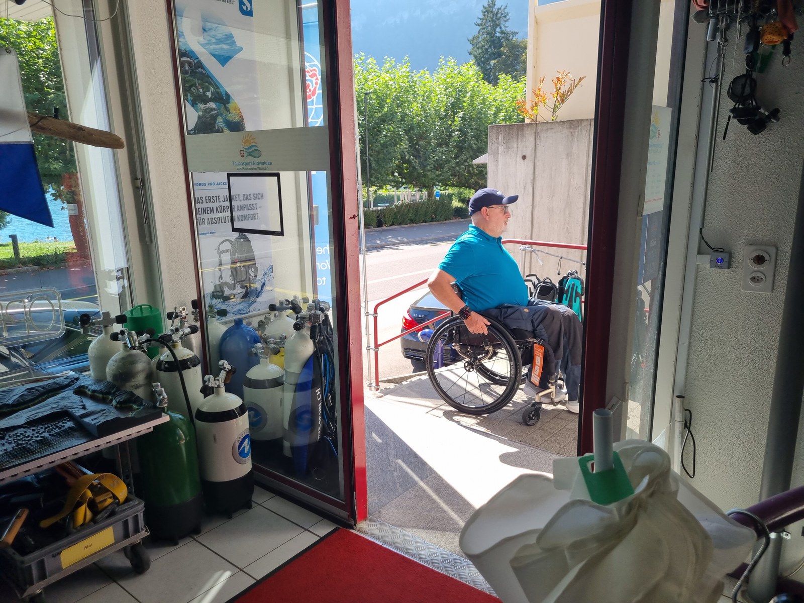Ein Mann im Rollstuhl vor der offenen Ladentür von Tauchsport Nidwalden in Hergiswil, drinnen Tauchflaschen.
