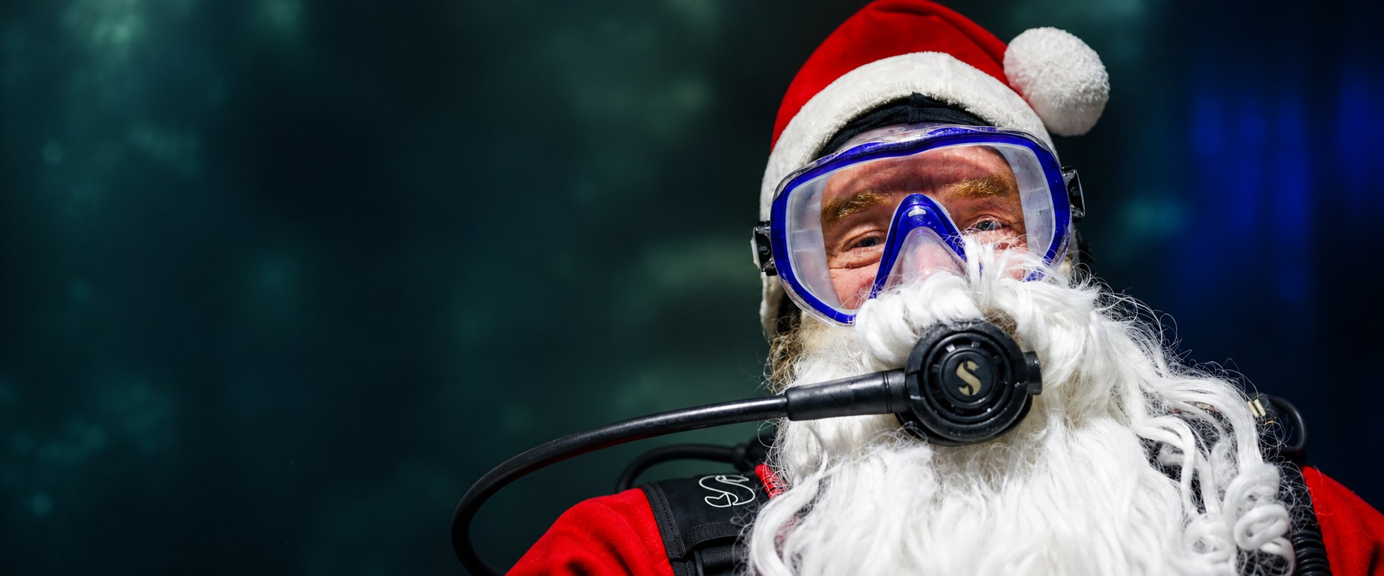 Als Samichlaus verkleideter Taucher mit weissem Bart, roter Mütze, Taucherbrille und Atemregler vor dunklem, unscharfem Wasser