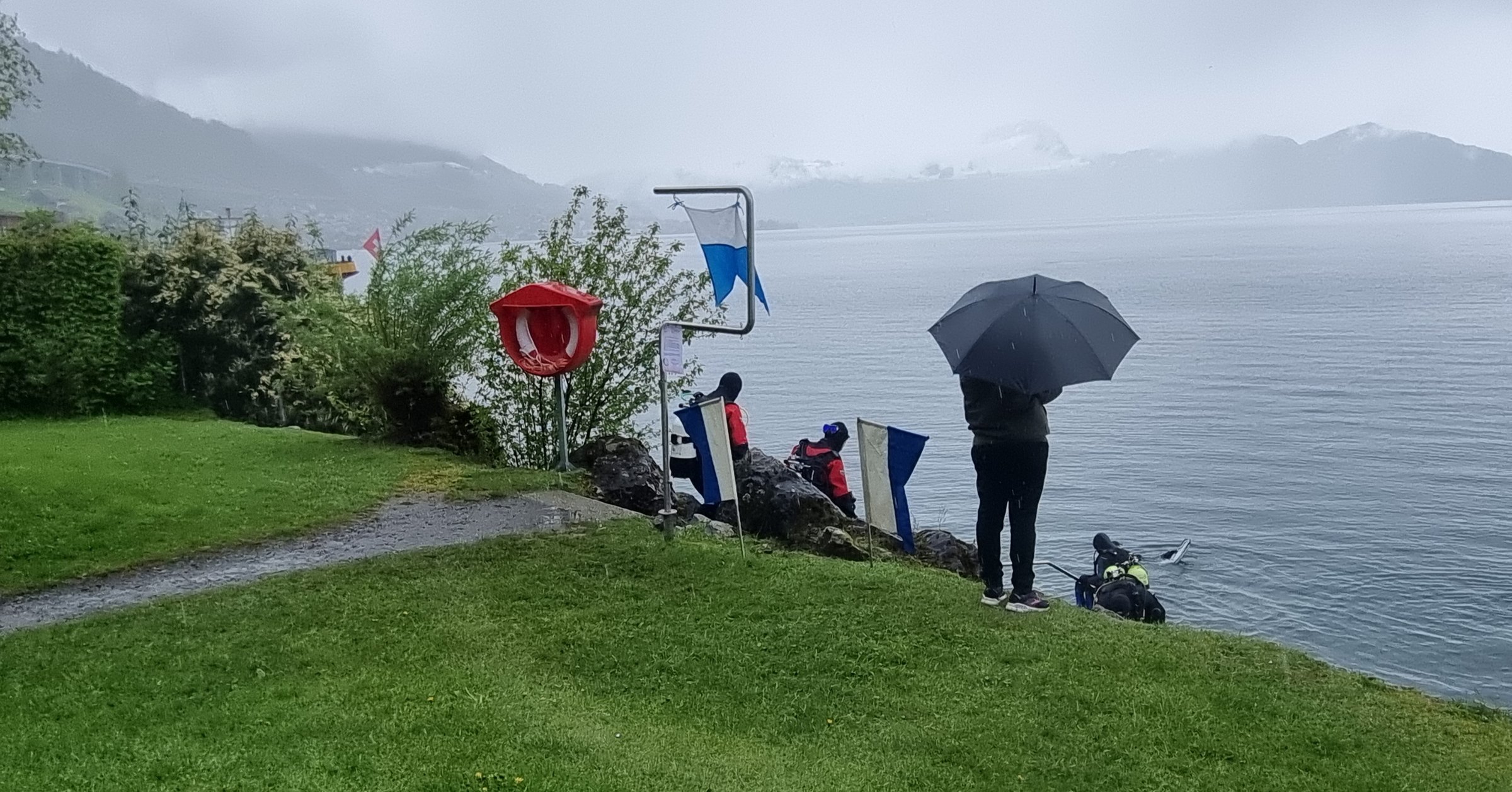 Taucher steigen bei Regen in den Vierwaldstättersee, am Ufer steht eine Person mit einem Regenschirm.