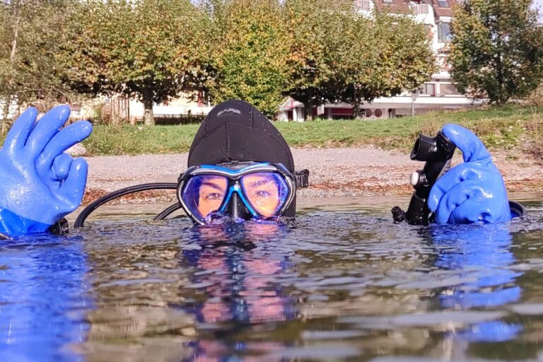 Taucherin schaut mit Maske knapp über die Wasseroberfläche und zeigt mit blauem Handschuh das Okay-Zeichen